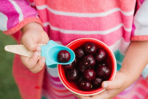 Ребенок держит мисочку с неочищенными черешнями и ложечку, в которой уже лежит одна ягода