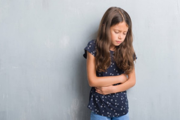 Грустная девочка стоит возле стены и держится за живот