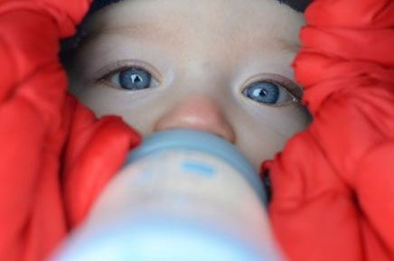 Укутанный грудничок пьет с бутылочки зимой, находясь в коляске