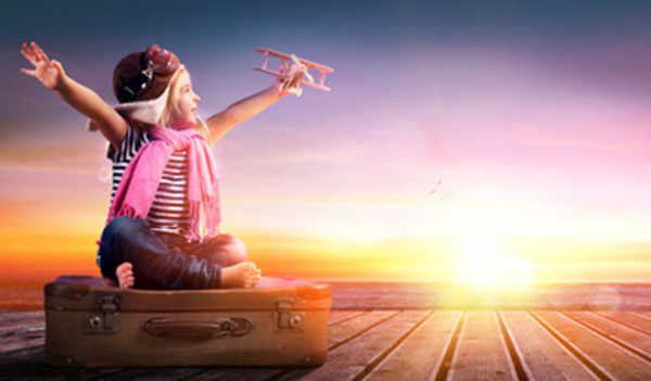 Ребенок в шапочке пилота сидит на чемодане и держит самолетик в руке