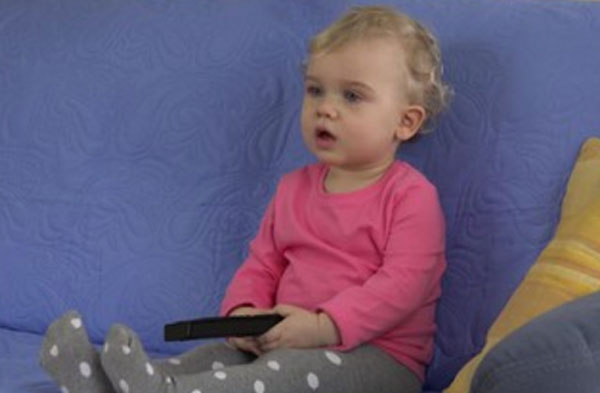 Годовалая девочка сидит с пультом от телевизора и смотрит прямо (по всей видимости на экран)
