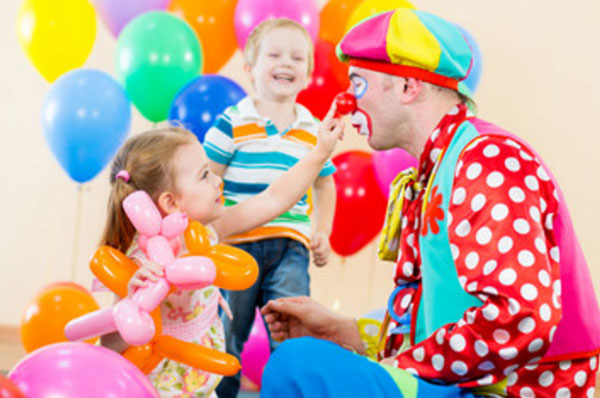 Клоун сидит возле девочки, которая пробует потрогать его нос. На заднем плане счастливый мальчик и воздушные шары