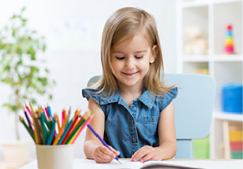 Девочка ровно сидит за столом и рисует карандашами