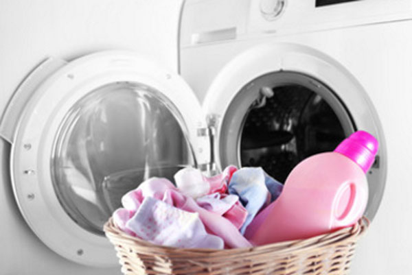 Открытая дверца стиральной машинки и корзинка с детской одеждой и гелем для стирки