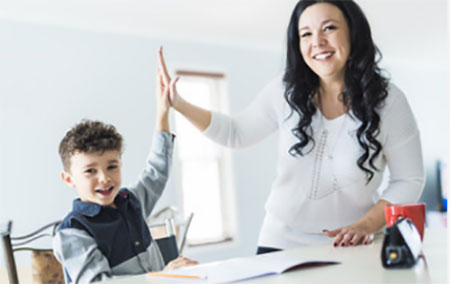 Мама хлопает одной рукой в ладош своего сына. Оба рады. Ребенок сидит за столом, перед ним открытая тетрадь