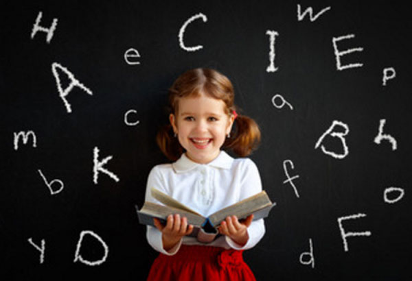Девочка младшего школьного возраста стоит с открытой книгой в руках. Сзади нее школьная доска, на которой мелом написаны английские литеры
