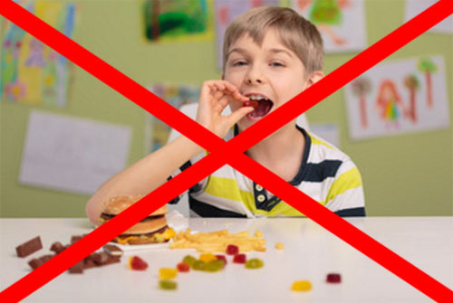 Ребенок ест сладости, перед ним лежат конфеты, в том числе и шоколадные, гамбургер. Фотография перечеркнута