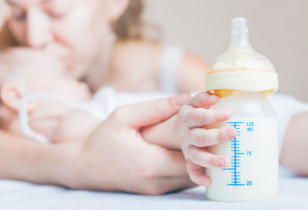 На переднем плане ручка грудничка с бутылочкой с молоком и смесью. На заднем мама, целующая карапуза в лобик
