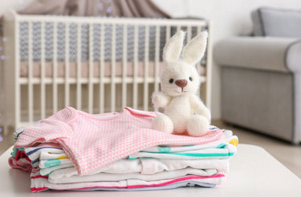 На заднем плане детская кроватка. На переднем - стопка одежды и мягкий заяц