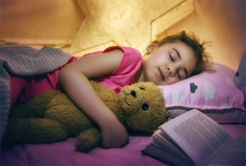 Девочка спит ночью в обнимку с плюшевым медведем. Рядом лежит раскрытая книга. Комната слегка освещена