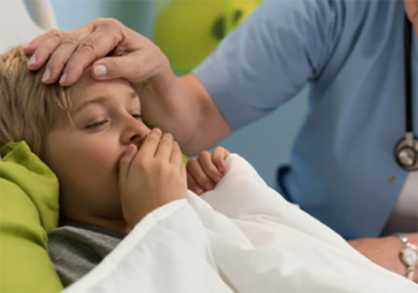 Ребенок лежит в постели, кашляет. Доктор держит руку на его лбу