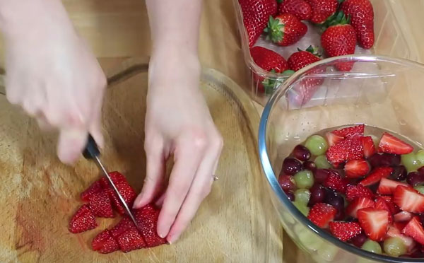 В одной емкости вымытая клубника, в другой нарезанный виноград и клубника, которые плавают в сиропе. На досточке продолжают нарезать