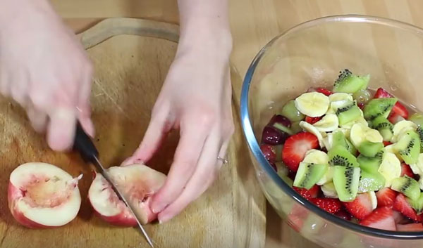 Разрезанный напополам персик собираются нарезать. Рядом емкость с другими фруктами