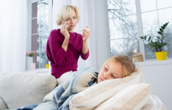 Девочка лежит в постели. Мама стоит сзади дивана и смотрит на градусник