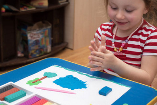 Девочка скатывает пластилин в ладошках. Перед ней лежит картина из пластилина