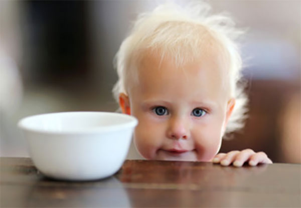 Годовалый мальчик стоит возле стола, на котором стоит миска