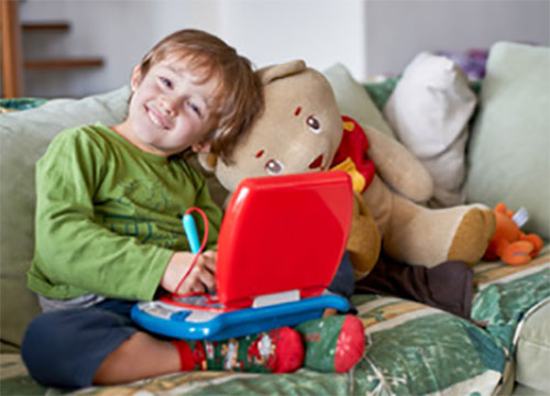 Ребенок улыбается. Сидит на диване и занимается на детском ноутбуке, рядом плюшевый медведь